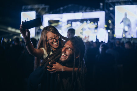 Teenage couple at summer music festival taking selfie with a smartphone while dancing in front of...