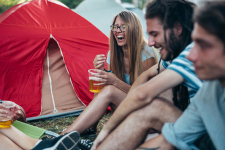 Teenagers sitting on the ground in front of tents on a music festival. Happy friends playing music and drinking beer in camping party. Friendship.の写真素材
