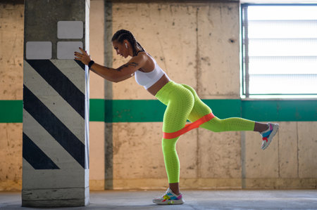 Young attractive woman training outside while using resistance band with a concrete wall in the...