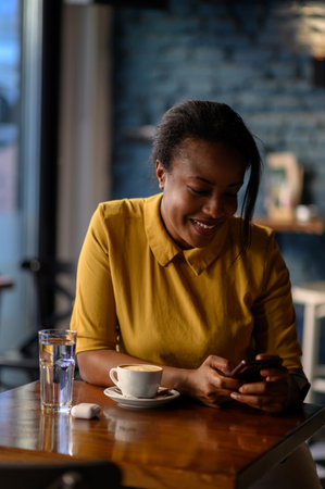 Beautiful young african american woman using a smartphone and drinking coffee while sitting in a cafeの写真素材