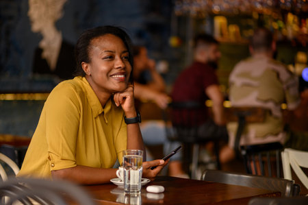 Beautiful young african american woman using a smartphone and drinking coffee while sitting in a cafeの写真素材