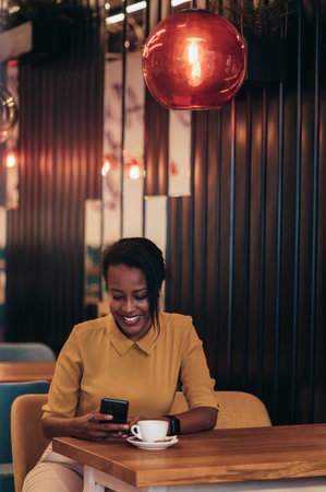 Beautiful young african american woman using a smartphone and drinking coffee while sitting in a cafeの写真素材
