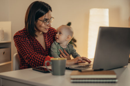 Beautiful business mom using a laptop and smiling while spending time with her cute baby boy at homeの写真素材