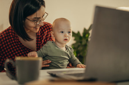 Beautiful business mom using a laptop and smiling while spending time with her cute baby boy at homeの写真素材