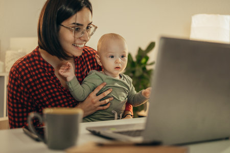 Beautiful business mom using a laptop and smiling while spending time with her cute baby boy at homeの写真素材