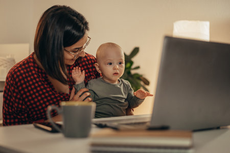 Beautiful business mom using a laptop and smiling while spending time with her cute baby boy at homeの写真素材