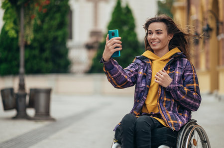 Young beautiful woman with disability who uses a wheelchair taking a selfie with her smartphone...