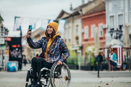 Young beautiful woman with disability who uses a wheelchair taking a selfie with her smartphone while out in the cityの写真素材