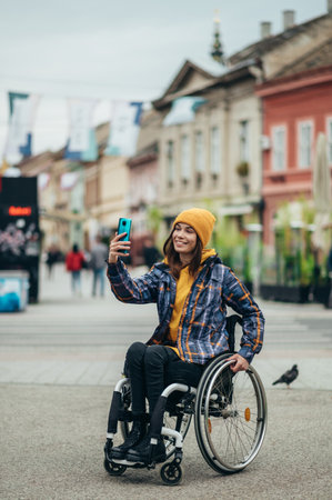 Young beautiful woman with disability who uses a wheelchair taking a selfie with her smartphone while out in the cityの写真素材
