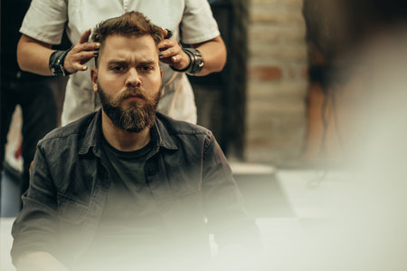 Young bearded man getting talking with his hairdresser while sitting in chair at barbershopの写真素材