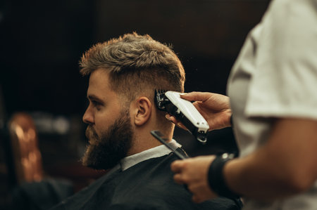 Young bearded man getting haircut by hairdresser while sitting in chair at barbershopの写真素材