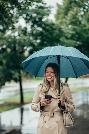 Young smiling businesswoman with umbrella and shoulder bag using smartphone and walking down city street during rainの写真素材