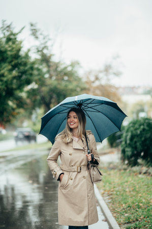 Young smiling businesswoman with umbrella and shoulder bag walking down city street during rainの写真素材
