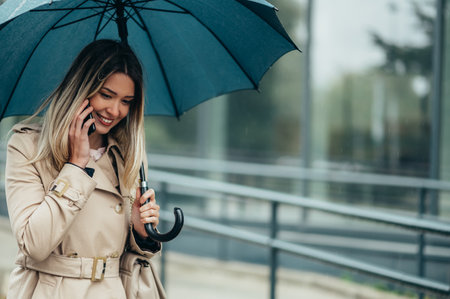 Young smiling businesswoman with umbrella and shoulder bag using smartphone and walking down city street during rainの写真素材