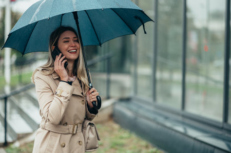 Young smiling businesswoman with umbrella and shoulder bag using smartphone and walking down city street during rainの写真素材