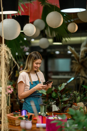 Florist using smartphone while standing next to the counter surrounded with flowers and plants in a flower shop