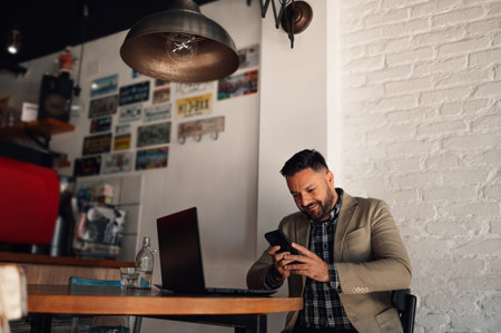 Handsome businessman using a smartphone and working on his laptop while drinking coffee in a cafeの写真素材