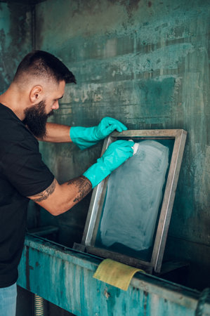 Male worker cleaning screen frame with a cloth in a printing workshop