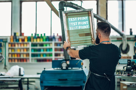 Male worker preparing screen printing film and wearing a dirty apron while working in a workshopの写真素材