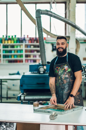 Male worker preparing screen printing film and wearing a dirty apron while working in a workshop