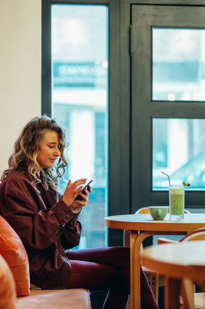 Beautiful young woman drinking matcha latte coffee and using smartphone in a cafeの写真素材