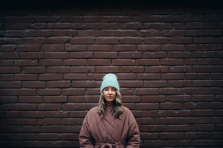 Beautiful young stylish woman wearing coat and a cap posing in front of a brick wall