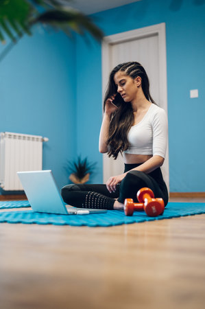 Gorgeous woman using laptop and a smartphone while training at home sitting on a fitness matの写真素材