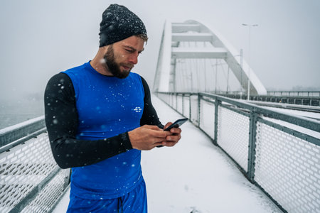 Young sportsman taking a break from running outside during snow and using smartphone. Healthy lifestyle. Attractive male running and training on a cold winter day.の写真素材