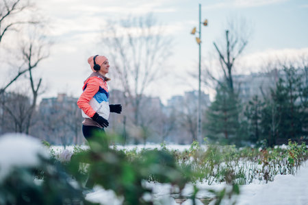 Happy young woman jogging in the park with snow on cold winter day and listening music on a headphones. Motivation. Urban city jogger. Training during a bad weather, snow and winter. Copy space.の写真素材