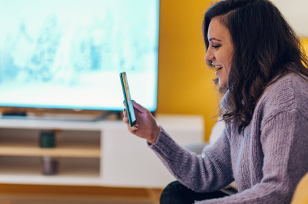 Smiling young hispanic woman sit on sofa at home and using a smartphone for a video call. Digital...