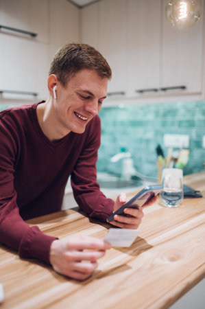 Joyful redhead positive man using a smartphone and smiling while looking at his credit card in the kitchen at home. Electronic payment. Online banking. Online shopping.の写真素材