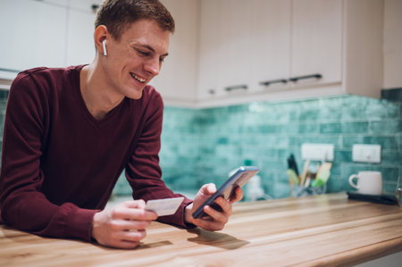 Young millennial redhead guy stand in the kitchen and using a smartphone while preparing to order food products delivery. Online banking. Online shoppingの写真素材