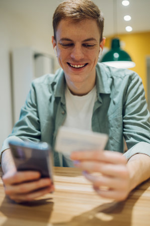 Handsome redhead man standing in the kitchen at home and using credit card to pay online with...