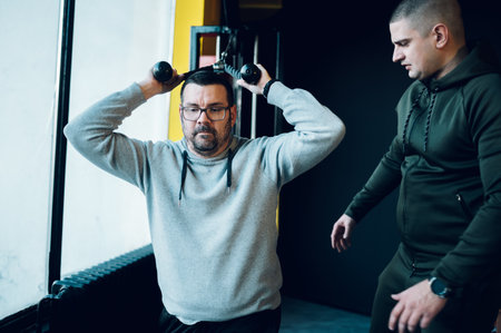 Obese middle aged men training in the gym with the help of a personal instructor and using a cross...