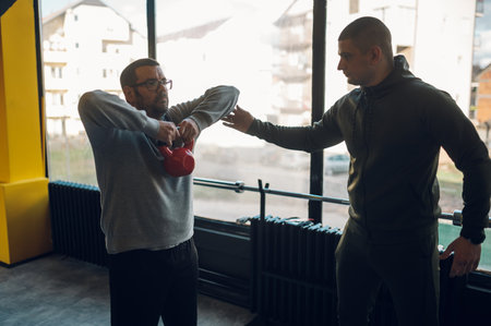 Obese middle aged men training with fitness trainer and using kettlebell in the gym. Overweight man trying to lose weight. Warming up.の写真素材