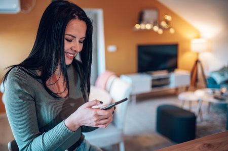Beautiful middle aged woman sitting at the kitchen counter while drinking morning coffee and using a smartphone at home. Leisure at home concept.の写真素材