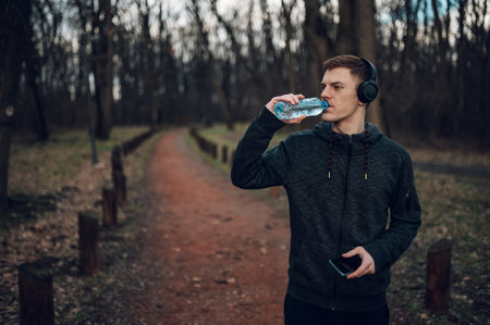 Redhead runner athlete taking a break from his workout and drinking water and using smartphone while...