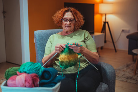 Senior woman sitting on an armchair at home and holding knitting needles and yarn knits clothes for...