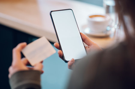 Woman hands holding credit card for shopping online and a smartphone while in coffee shop. White mockup smartphone screen. Online payment concept.Focus on a smartphone screen. Copy space.の写真素材