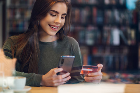 Young beautiful woman using smartphone and a credit card for online shopping while in a cafe during...