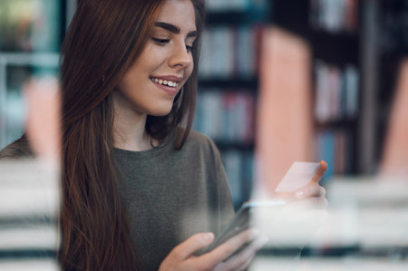 Young beautiful woman using smartphone and a credit card for online shopping while in a cafe during a day. Shopping online, booking tickets, making payment. Viewed through a window of a cafe.の写真素材