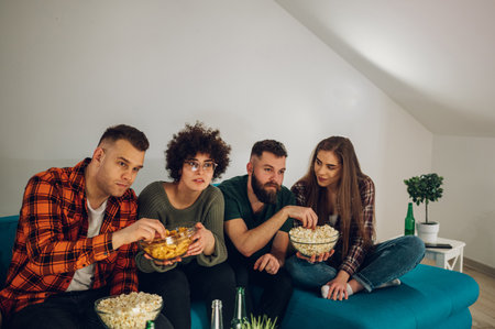 Friends relaxing together at home and watching television and eating snacks. Watching interesting movie at home. Spending weekend together at home.の写真素材