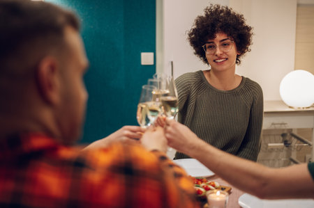 Beautiful young people enjoying meal while sitting at the dinning table in the kitchen together. Friends gathering together at home for dinner while toasting.の写真素材