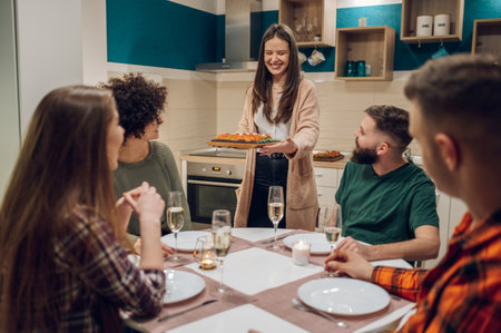 Group of cheerful young people enjoying dinner while sitting at the kitchen table together. Having a great time with friends during dinner while drinking and having funの写真素材