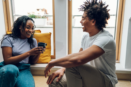 Cheerful african american couple drinking coffee or tea in the morning while sitting on a windowsill by the window at home. Romantic morning at home.の写真素材