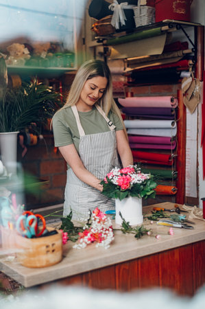 Young blonde florist woman creating flower arrangement in a round box for a gift for a special occasion while working in a flower shop. Beautiful bouquet of mixed flowers. View through the window.の写真素材