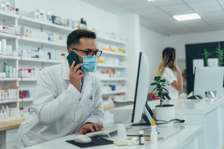 Male pharmacist with protective mask on his face using a smartphone while working at a pharmacyの写真素材