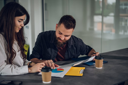 Two happy young businesspeople working together and discussing charts and sales reports and having a meeting in the open space office. Two successful young entrepreneurs collaborating on a new project.の写真素材