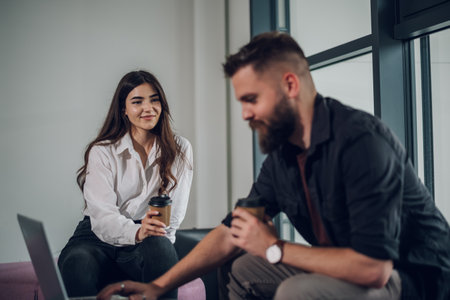 Portrait of a two young businesspeople using a laptop and drinking coffee takeaway while sitting in the office. Two business colleagues having a discussion during a meeting. Focus on a caucasian womanの写真素材