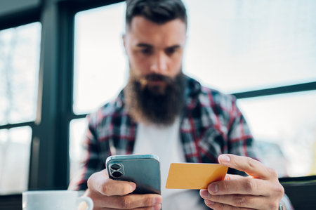 Young male in checkered shirt with long beard making transaction while using banking application on...